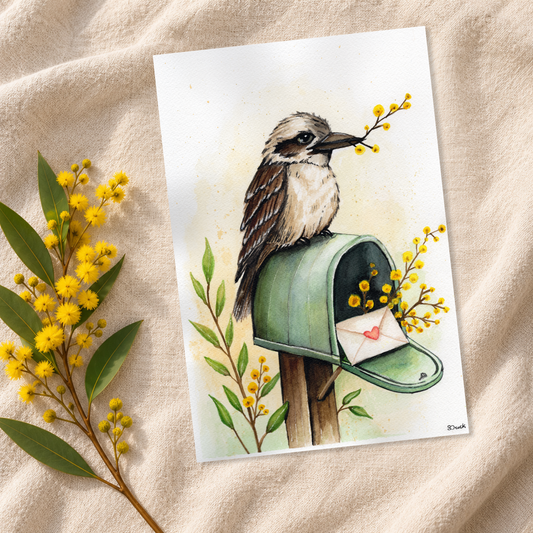 A watercolor art print featuring a kookaburra perched on a green mailbox holding a branch with yellow flowers, styled on a beige fabric background with yellow mimosa flowers.
